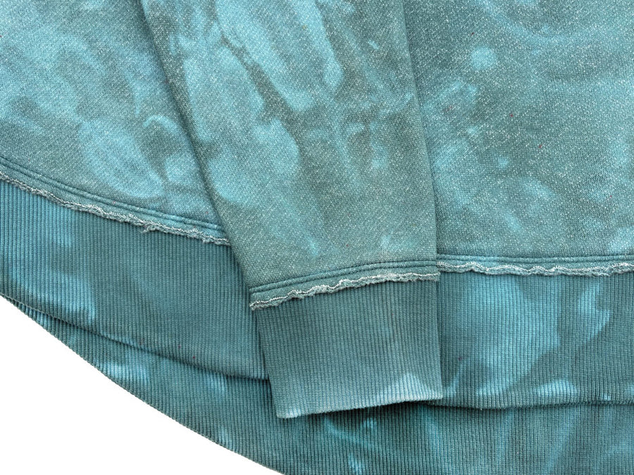 Dyed example: Close-up of teal sweatshirt fabric with a subtle organic dye pattern, and ribbed hems on a white background