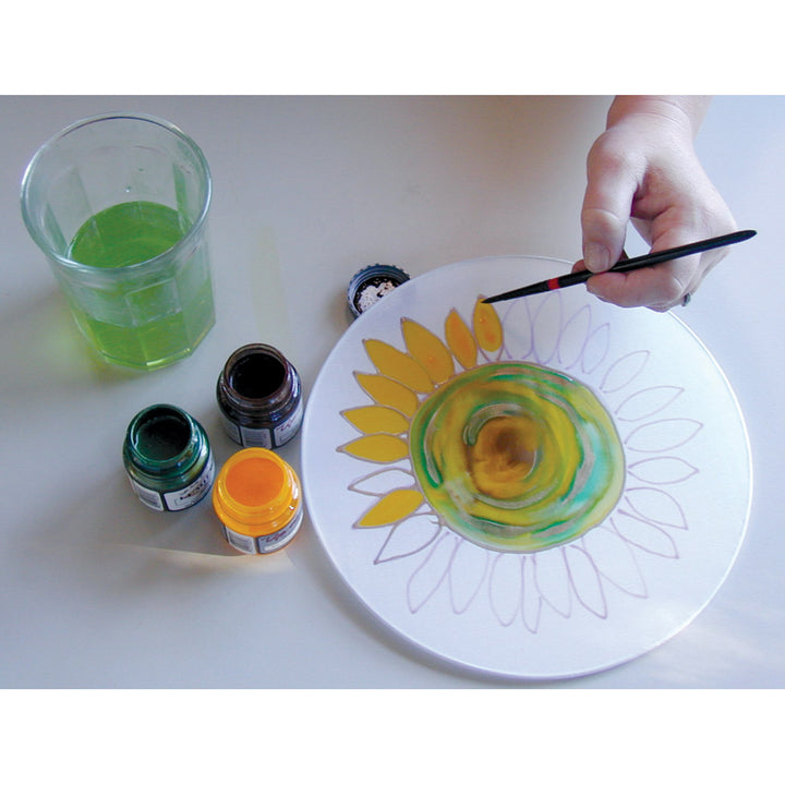 Hand with brush painting a silk hoop with a flower design with silk paint bottles on a white table.