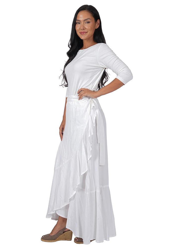 Left side view of a woman wearing a white top with an ankle length, side-tied, flowing white skirt on a white background.
