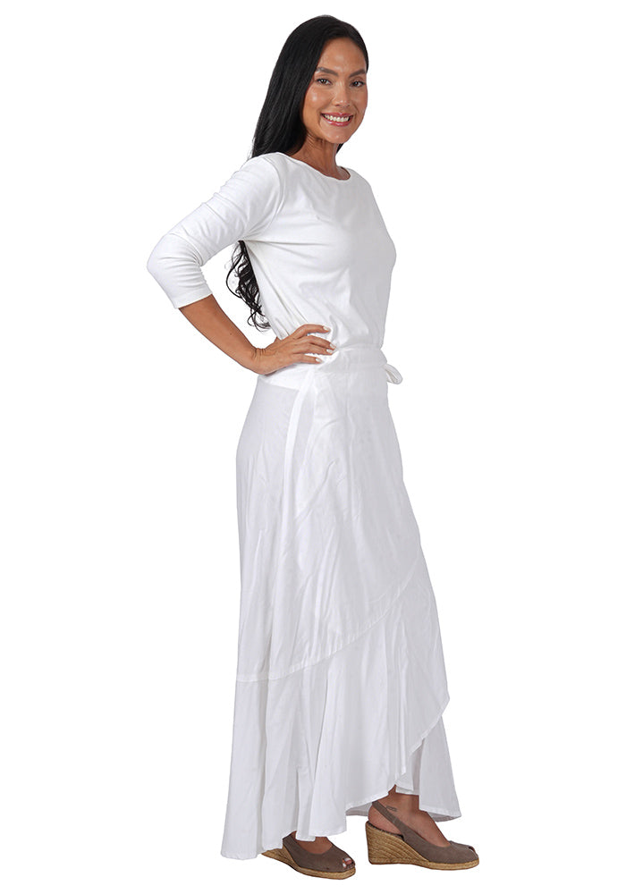 Right side view of a woman wearing a white top with an ankle length, side-tied, flowing white skirt on a white background.