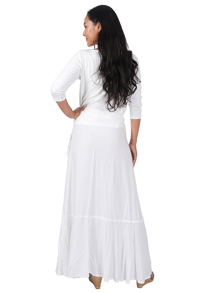 Back view of a woman wearing a white top with an ankle length, side-tied, flowing white skirt on a white background.