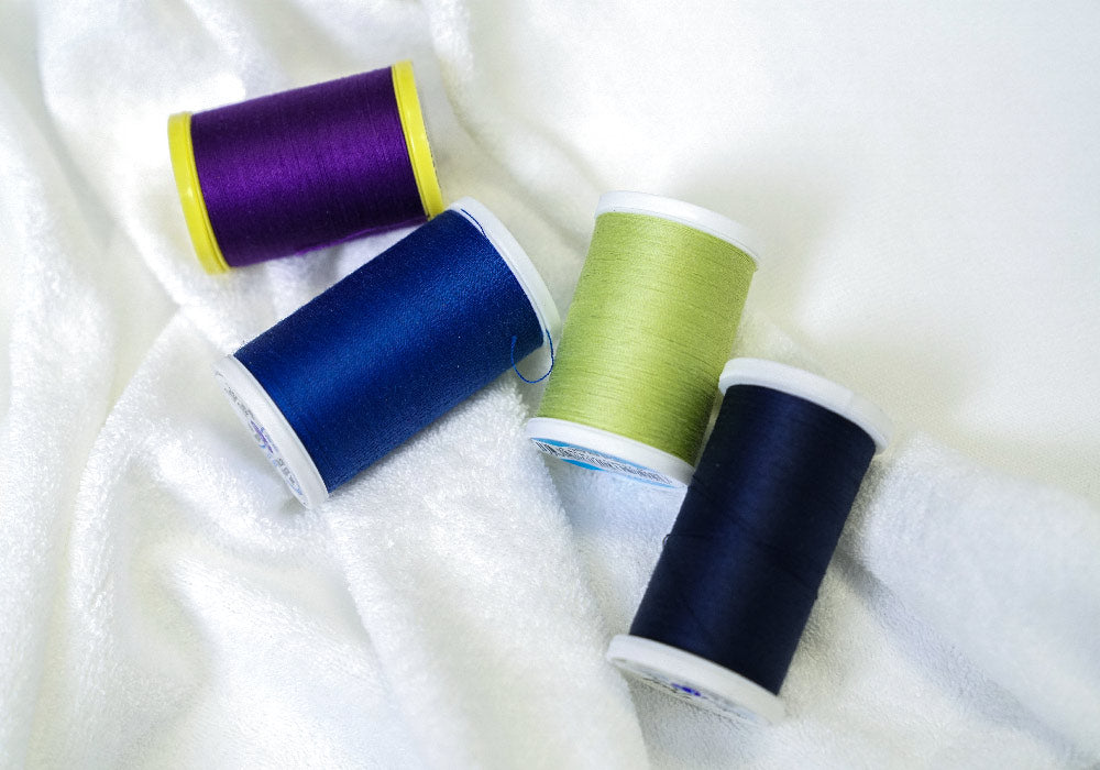 Four spools of thread in purple, blue, green, and navy on a white terry cloth fabric background