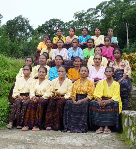 Group of Indonesian women in colorful traditional clothes, sitting together with a forest in the background.